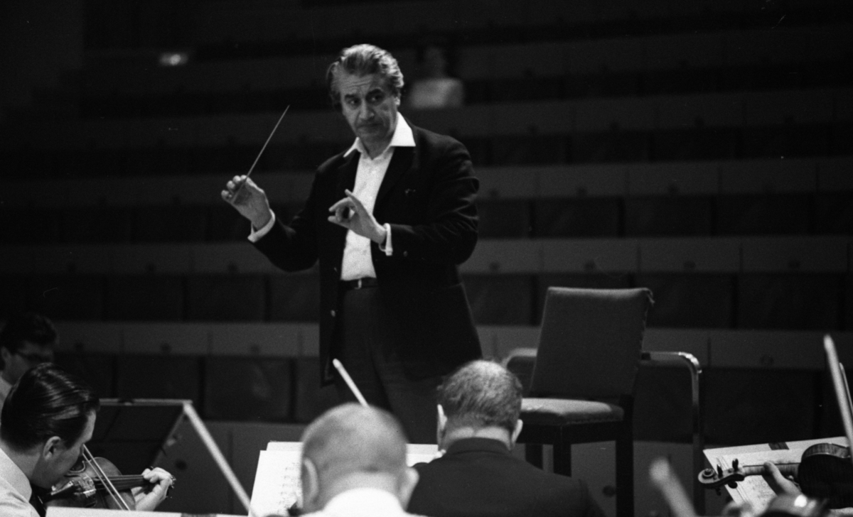 Cellibedache 13 juni 1966En dirigent klädd i vit skjorta och svart kavaj viftar med taktpinnen framför en orkester. Framför honom sitter musiker och spelar.