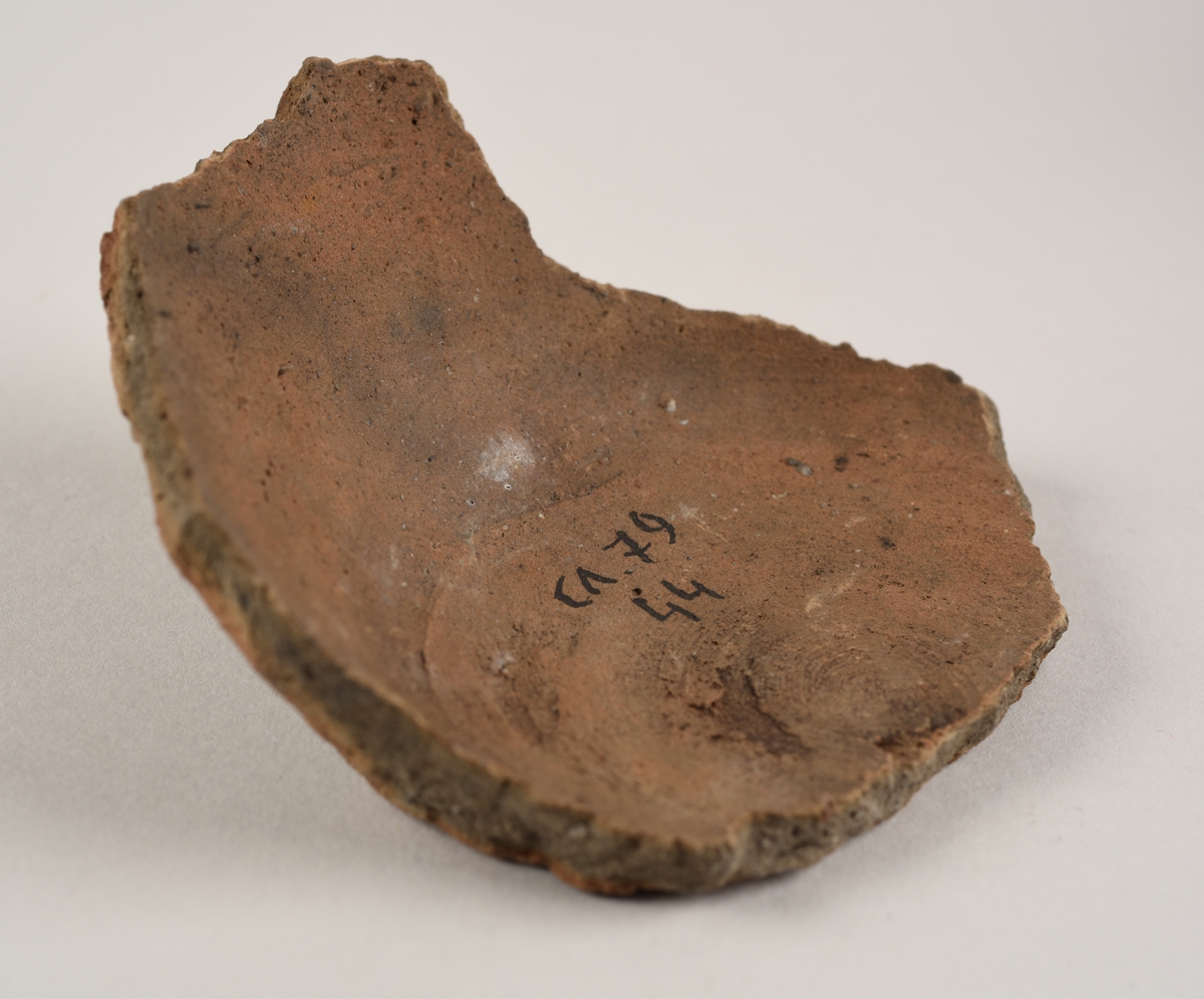 Keramikkfragment med en buet vegg og flat bunn. Fragmentet har bruddkanter og er mørkebrunt på utsiden og rødlig på innsiden.