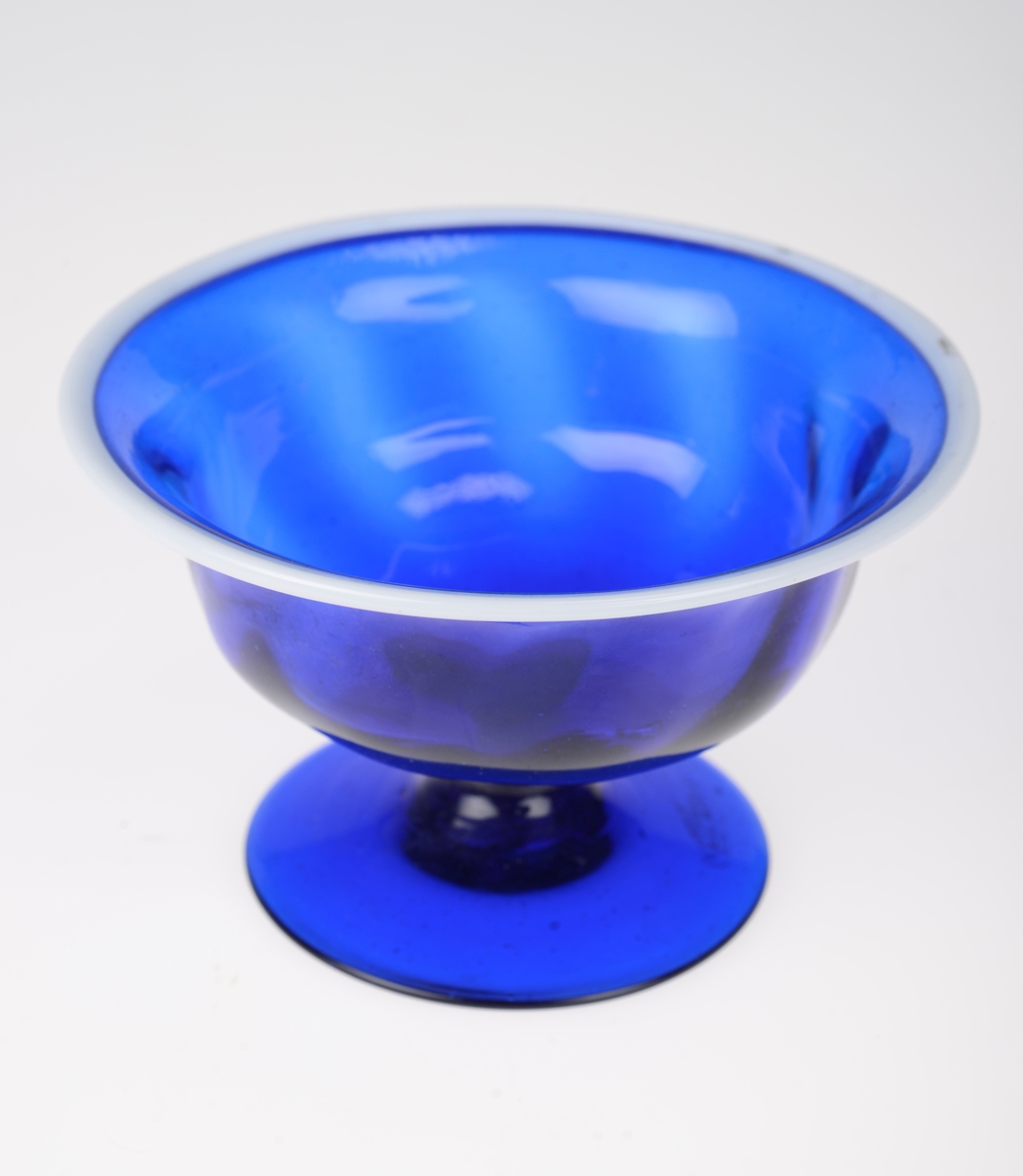 Mørk blå stettskål i glass. Bollen har en svak dekorering i form av lysere mørk blå skråstilte striper. Randen på skålen er i hvitt glass. Det kan sees små luftbobler i det blå glasset.