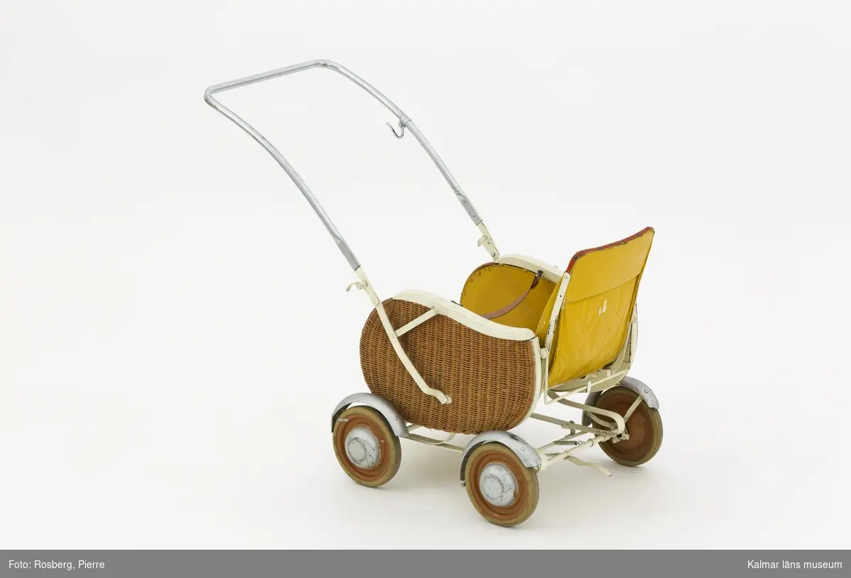 KLM 28596:2. Barnvagn, sittvagn, metall, trä, färg, textil, läder. Sittvagn för barn, ihopfällbar med stomme av järn och fyra hjul och reglerbart fotstöd av trä och järn. Sittkorgen har fastspikad klädsel av textil, målad med gul färg, och rödmålat, stoppat nackstöd. Ryggstödet är reglerbart i två lägen. Tvärs över sitsen hänger ett uppspänt läderband som fästs med hyska för barnets säkerhet. Ihopfällbart och reglerbart handtag av järn, övre delen kromat, och en krok på vänster sida. Vagnens sidor har ett korgflätat yttre lager, vitmålade sargar och silvermålade stänkskärmar och navkapslar.