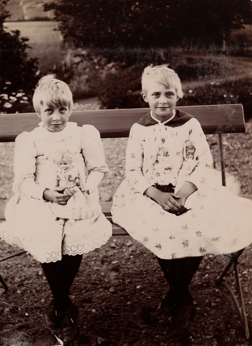 Eva og Minnie (Emilie) Christensen sitter på en benk med hver sin dukke i armen.
