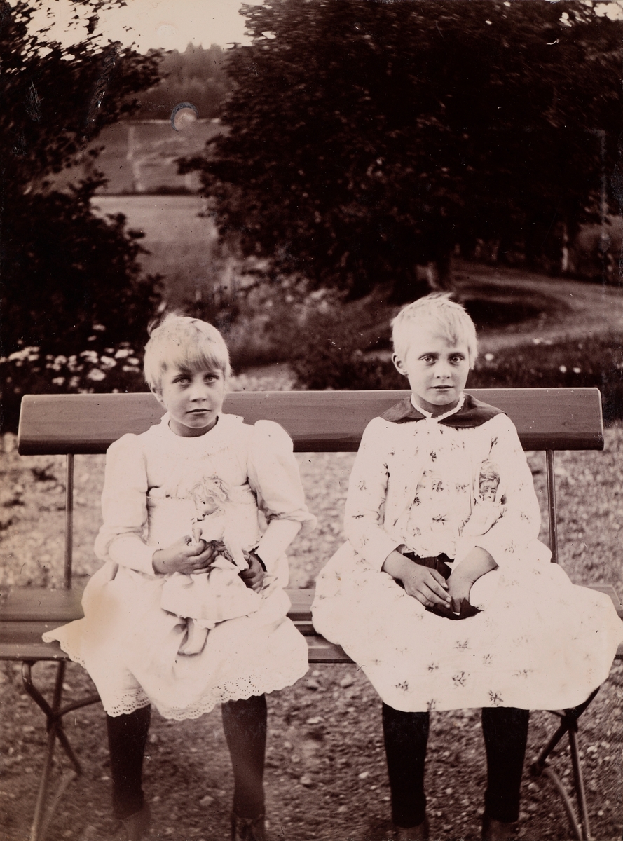 Eva og Minnie (Emilie) Christensen sitter på en benk med hver sin dukke i armen.