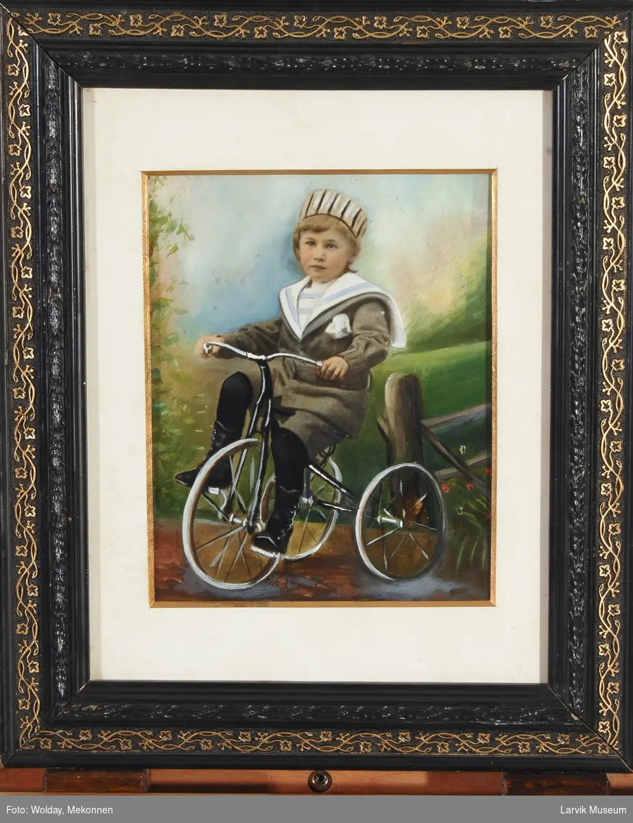 portrett av gutt som 3-4-åring på trehjul-sykkel iført strikkedress med matroskrave,hatt og svarte støvler