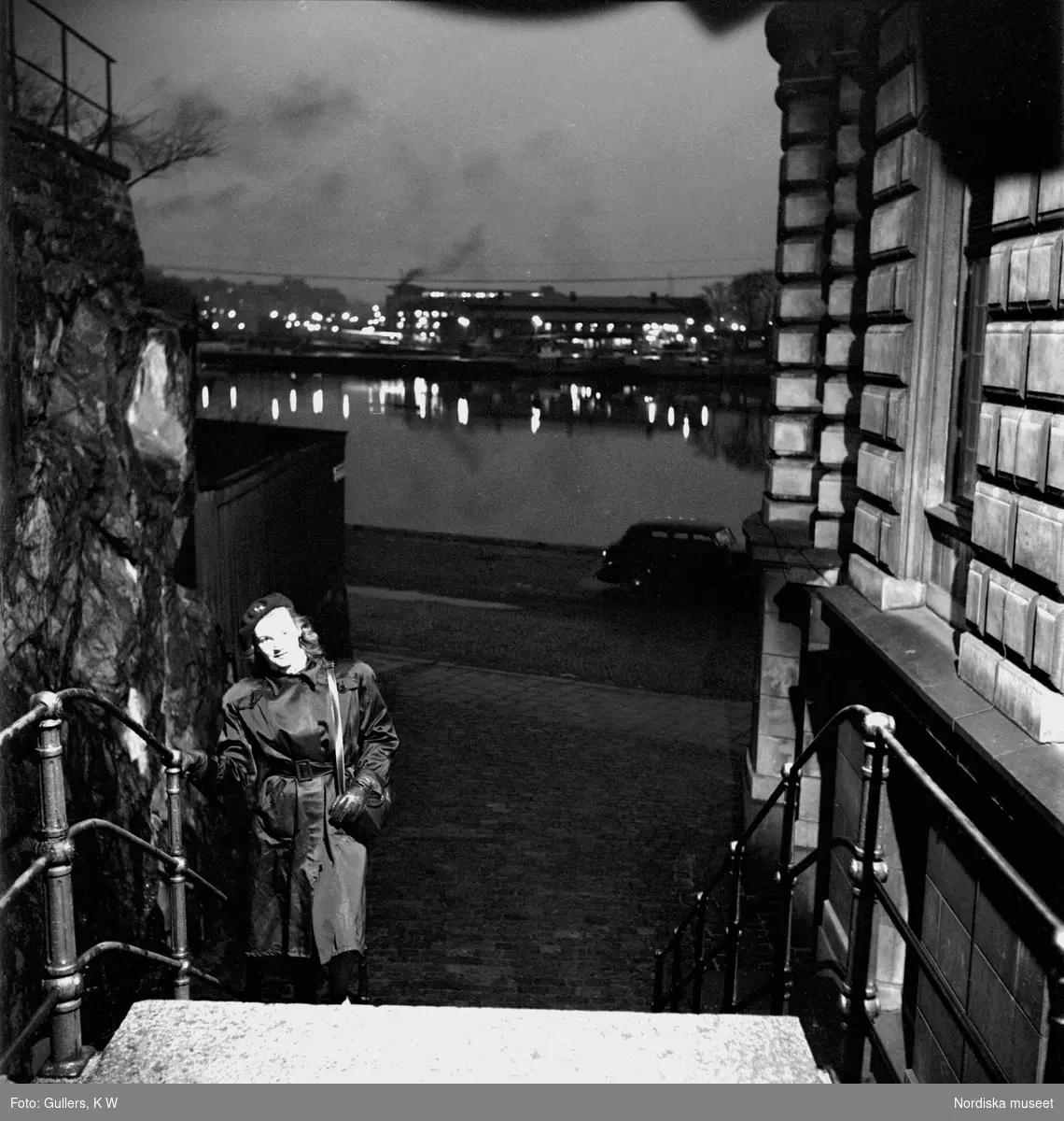 Stockholm. En ung kvinna går uppför trappa i förgrunden. Stadens ljus speglar sig i vattnet i bakgrunden.