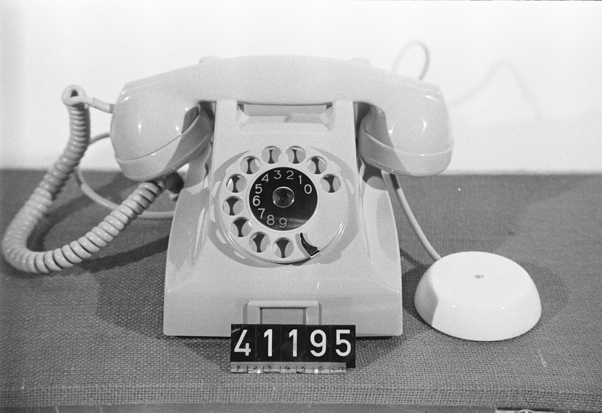 Telefonapparat BC 664, sidoapparat för AT-system. Bordapparat av grå plast modell m50 med apparatsnöre anslutet till väggplint med vitt plastlock.