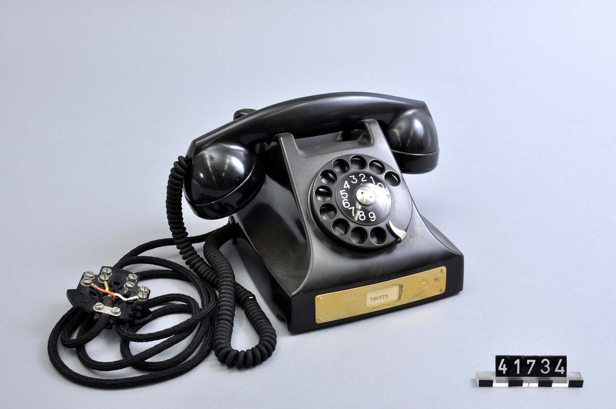 Telefonapparat BC 560, ändstationsapparat för AT-system. Bordtelefon modell m50 av svart bakelit med silverplåt och textilklätt apparatsnöre anslutet till väggplint utan lock.