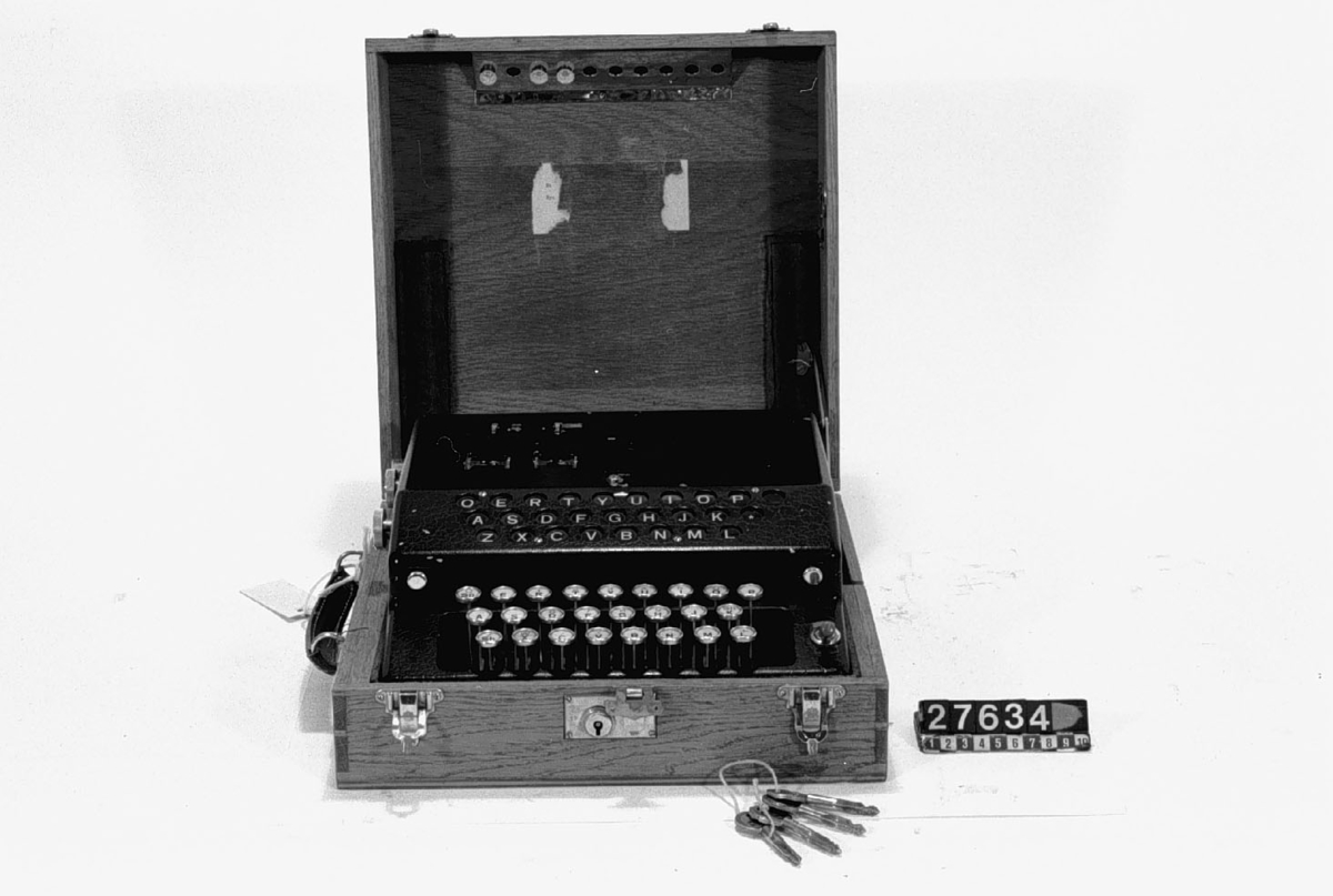 Mekanisk krypteringsmaskin med tangentbord, fyra inställningshjul och lamptablå för utdata. I träetui.
Tillbehör: Fyra st nycklar.