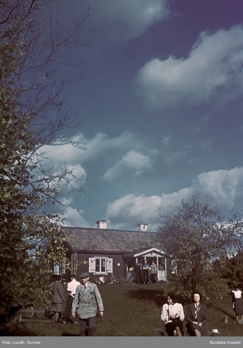 Sommarlandskap, på gräsplanen framför en parstuga med fönsterluckor rör sig människor. Två kvinnor sitter på en bänk framför ett frukträd i solen. Flera män och kvinnor sitter i gräset lutade mot husväggen och på och framför husets veranda har flera män samlats. En man promenerar bort från huset ut ur bild.
