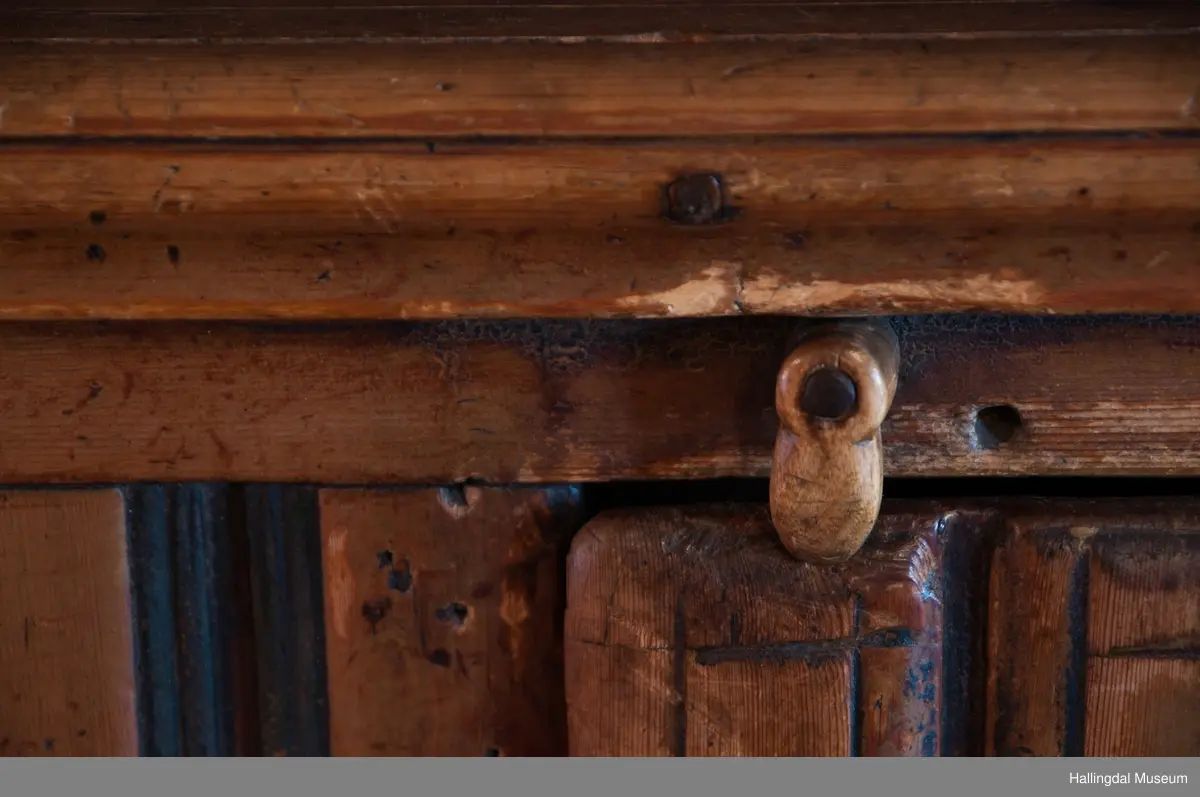 Rester etter rød og blå maling.  Gesims, skråner ned bakover. Profilert. 6 sirkulære hull i arkitraven.  Mellom disse er malt med sorte bokstaver.  Gavlene har utskårne "hylleknekter".  Bakveggen består av fire vertikale planker.  Skjøtene er dekket med profilerte lister.  Bare to biter i nederste del til venstre er fremdeles på plass.  Nederste del er lukket skap.  Tredelt, midterste del er skapdør.  Platen har profillist.  Feltene har speilfyllinger.  Blåmalte.  Klosset marmorering.  Speilfyllingenes form er som en liggen H.  Har profillist.  Skapet er satt på en ny grønmalt plate.  På døren er malt hvite bokstaver.  Nederst på døren er skåret inn bokstaver, opp-ned.  To nøkkelhull m. jernbeslag på venstre side av døren.  Over døren til venstre, en dreibar trekloss, holder igjen.  Innvendig har døren merke etter stort jernbeslag på samme side som nøkkelhullene.  To lister skåret i treverket minner om lange jernbeslag,-smalner mot nøkkelhullene.  Akantusranker i rødt, blått og hvitt, meget avslitt.  En hylle i skapet. Mangler bakvegg.  Karmen har på venstre side spor etter to jernhengsler.  Øverst på høyre kortside, mangler gesimsen en bit, og holdes til sideveggen med en jernkrampe.  Jernkrampe også på venstre side.  Skapplaten mangler list på høyre side.  Venstre side, fra gesimsen og ned har spor etter å ha stått inntil en vegg eller et annet møbel.