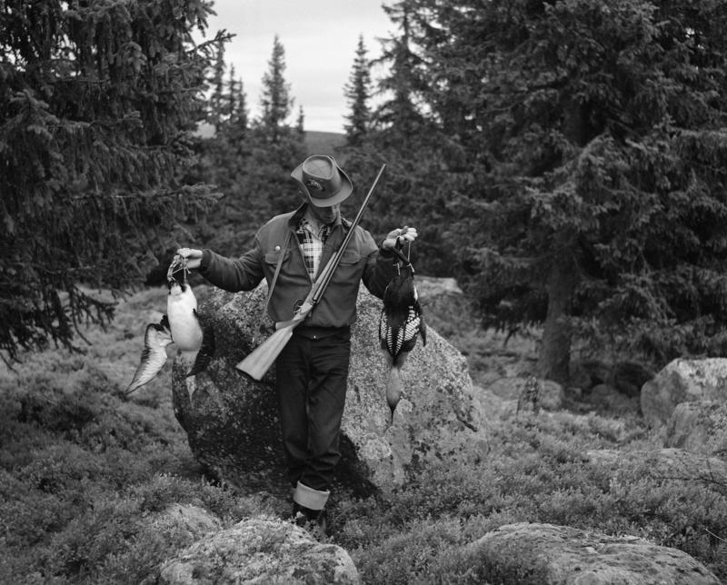 En jeger står i skogen med geveræt hengende i en reim over magen. I hver hånd holder han en rype.