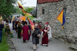 Gruppe mennesker med fargerike middelalderflær i rødt, grønt, gult, blått og hvitt som går parade. To middelalderfaner i gult og rødt.