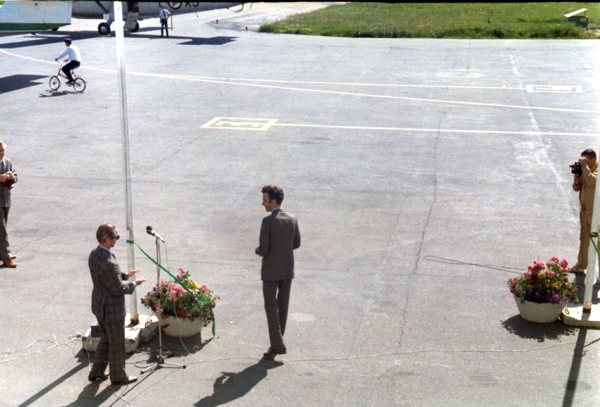 Lufthavn/Flyplass. To fly, DHC-6 Twin Otter fra Forsvaret og Widerøe skimtes. Midt på plassen stå to personer ved en mikrofon og blomsterdekorasjon. Fotografer knipser ivei.