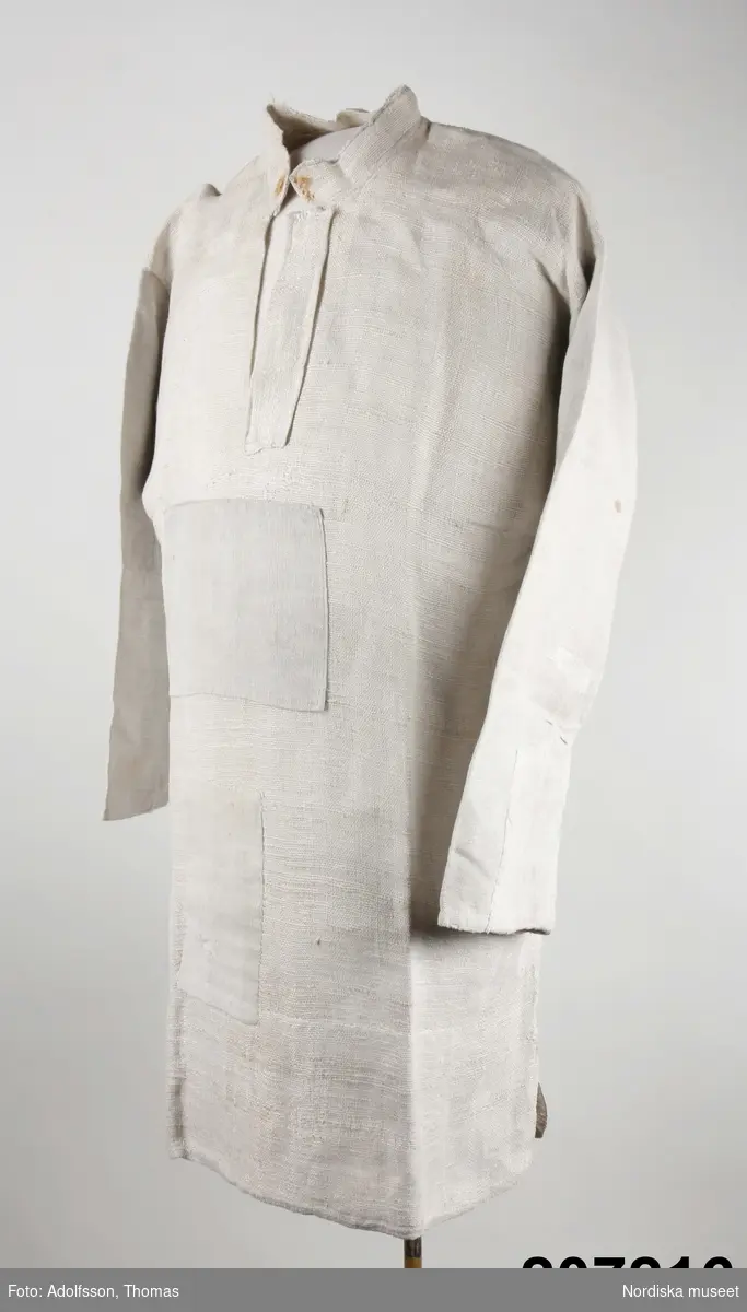 Långskjorta av vit tämligen grov och glesvävd linnelärft.  Ett bålstycke med sidsömmar och sidsprund, vidsydd rak ärm med ärmspjäll, ärmtyget  inte lika glest som båltyget.  2,5 cm hög ståndkrage med knapphål och rostfläck efter en knapp.  19 cm halssprund med  4 cm brett förslag Skjortan maskinsydd. 
Anm. Sliten och  lappad med finare  linnetyg  mitt fram, samt på ärmarna. Dessutom konserverad i museet med grovt linne på insidan
 Har brukats av hammarsmeden fredrik lejström f. 1842,  vid  Strömbacka bruk i Hälsingland. Smederna bar linneskjorta, träskor och benholkar som skydd mot värmen. Eftersom denna skjorta är kortare än  de brukar vara kan den vara senare när smederna börjat med långbyxor. Jfr med 207 224
/Berit Eldvik 2012-12-20
