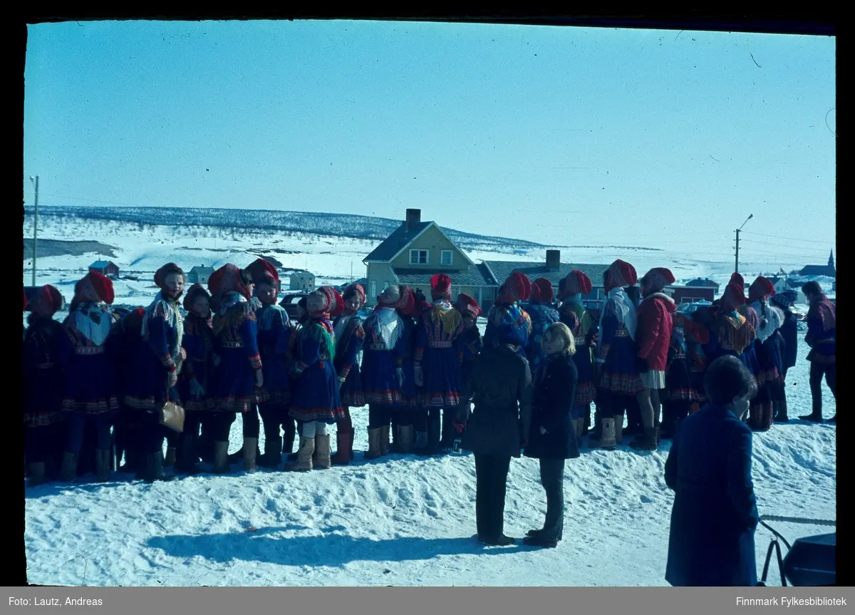 Kautokeino i 1969. Kronprinsparets besøk. (Harald og Sonja). 
Tilskuere kledd i samiske kofter, venter på et glimt av kronprinsparet.