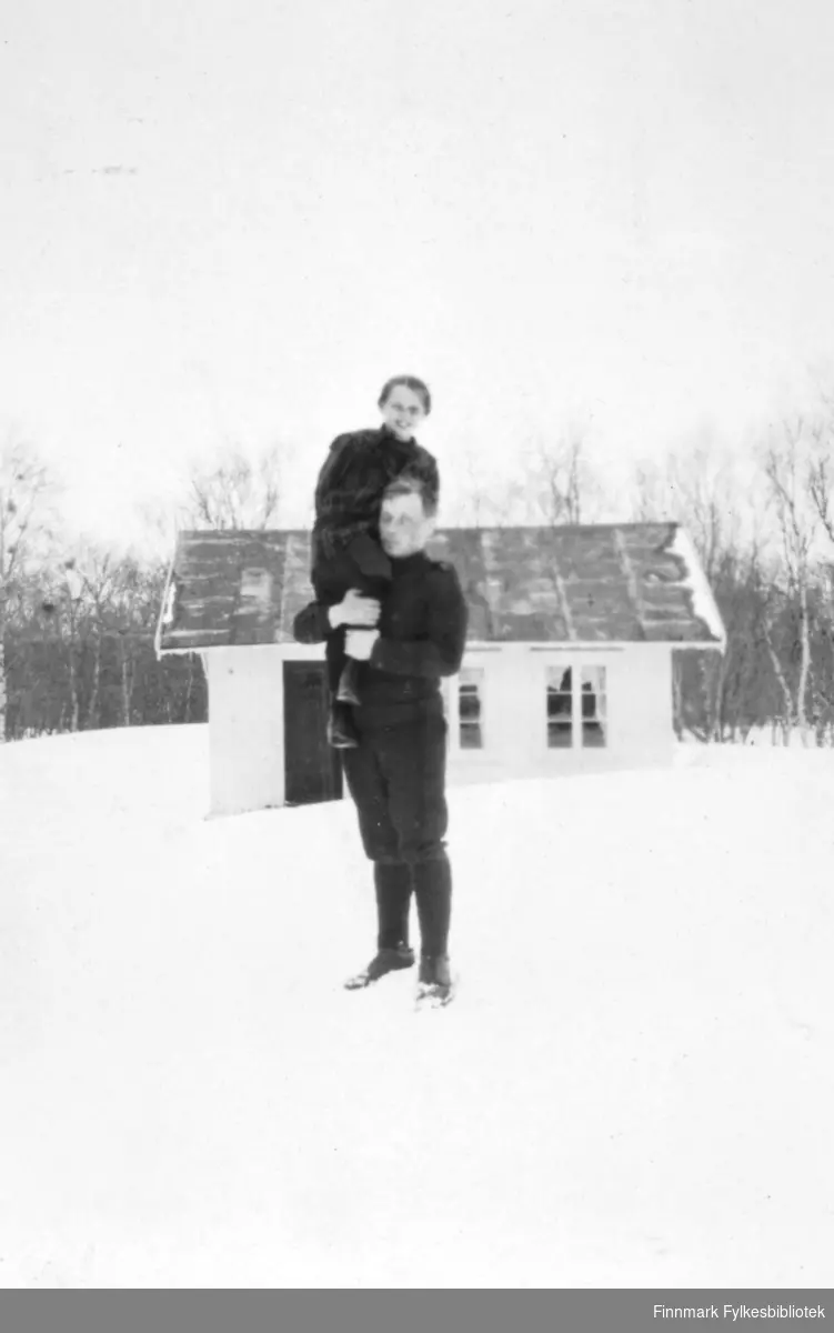 Kontorbygningen, Fredenslund, mai 1917. En mann står i snøen med en større gutt på skuldra. Mannen er kledd i genser med nikkers og strømper, gutten har en rutete vinterjakke på. I bakgrunnen ser vi en bygning, her beskrevet som kontorbygningen og er et relativt lite hus. I vinduene kan vi skimte gardiner og bak huset, krattskog. Fredenslund var Lensmann Strands gård, og lå i Bonakas, Tana. Bonakas, nordsamisk: Bonjákas, er en jordbruksbygd beliggende på vestsiden av Tanaelva i Tana kommune omkring ti kilometer ovenfor Tanamunningen.