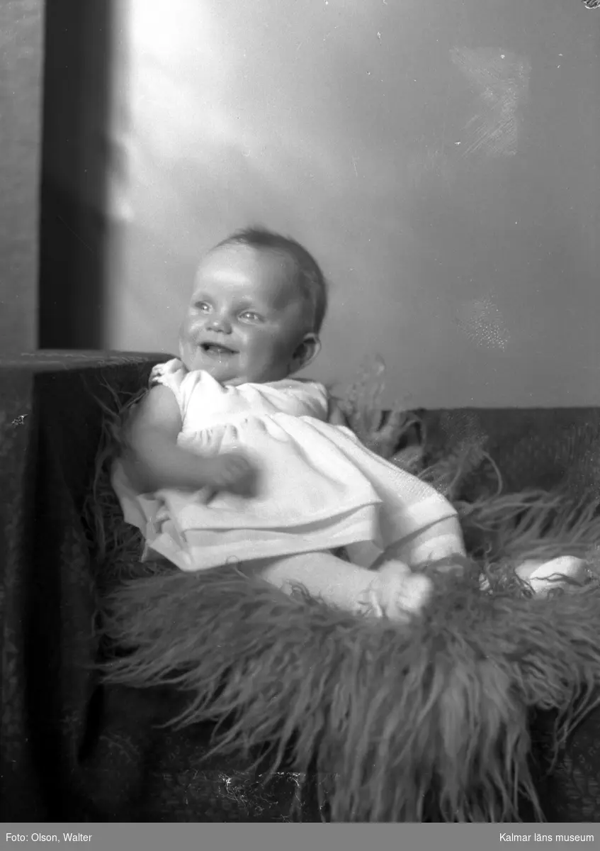 Ateljéfoto. Babyflicka sittande på fårskinnsfäll.