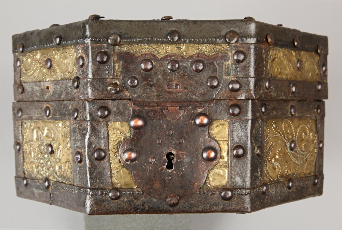 Åttekantet. Den har dekorerte flater i messing med jernbeslag og det hele er naglet på tre. På lokket er det en hank. Flaskefôret er låsbart, men den originale nøkkel mangler.