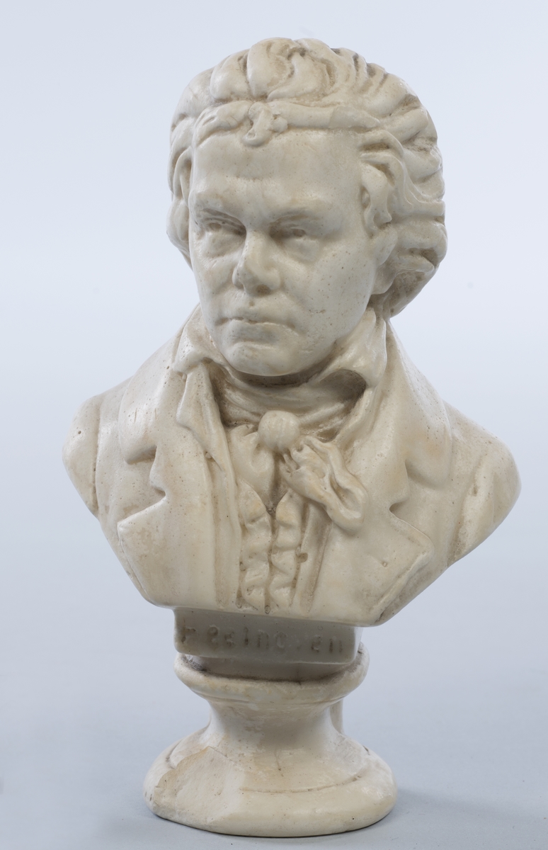 Beethoven iført åpen frakk, vest, skjorte med høy krage og knyttet tørkle i halsen. Rund sokkel.