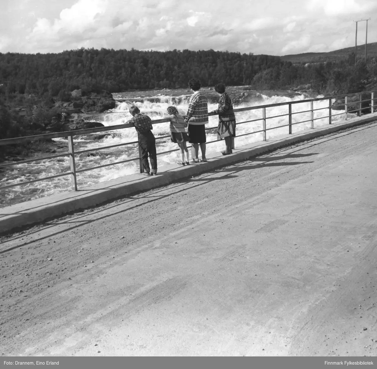 Jenny Drannem og Sonja Lappalainen ser mot Skoltefossen sammen med to barn Ole A. Gabrielsen og Kari E. Gabrielsen. Jenny og Sonja er tanter til Ole og Kari. De står på brua langs E6 som går over Neidenelva. Tidspunkt er 1960-tallet. Lignende foto FBib.06009-652.