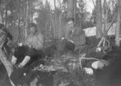 To menn sitter ved et lite bål i skogen. Sted og personer er