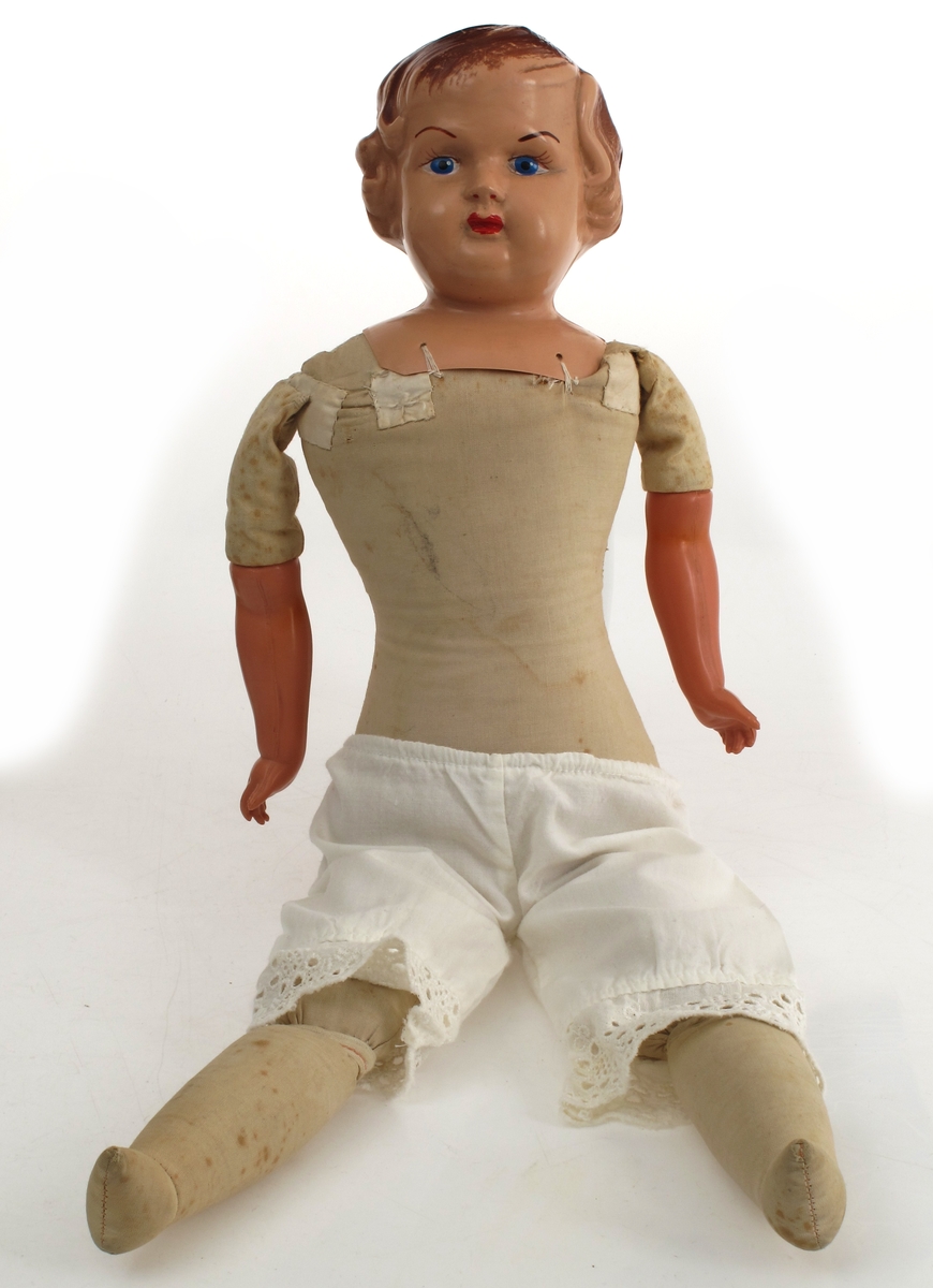 Dukke med dukkeklær. Hode og armer av celluloid, resten av dukken er tekstilt materiale. Kjole bomull, mørk blå farge, borelåslukking. Bukse mammelukker. 

Hodet er ikke merket av produsent, men har typisk KaySax-form.