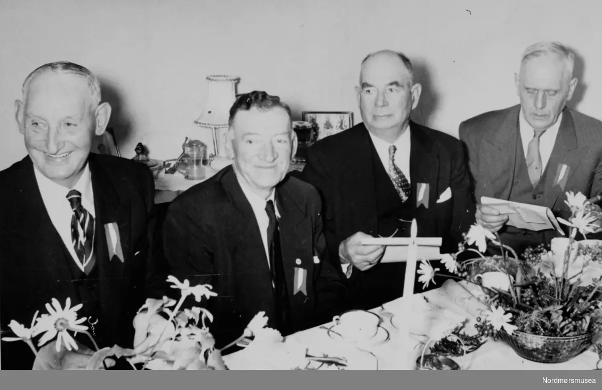Fire "gutter" sittende ved et pyntet kaffebord. Disse er Gustav Elgenes, Adolf Roesen, Erling Gaustad og Arne Holden. Fotoet er fra Vågens guttekorps jubileumsmøte hjemme hos Alfred Andersen den 18. oktober 1956. Fra Nordmøre Museums fotosamlinger. 

