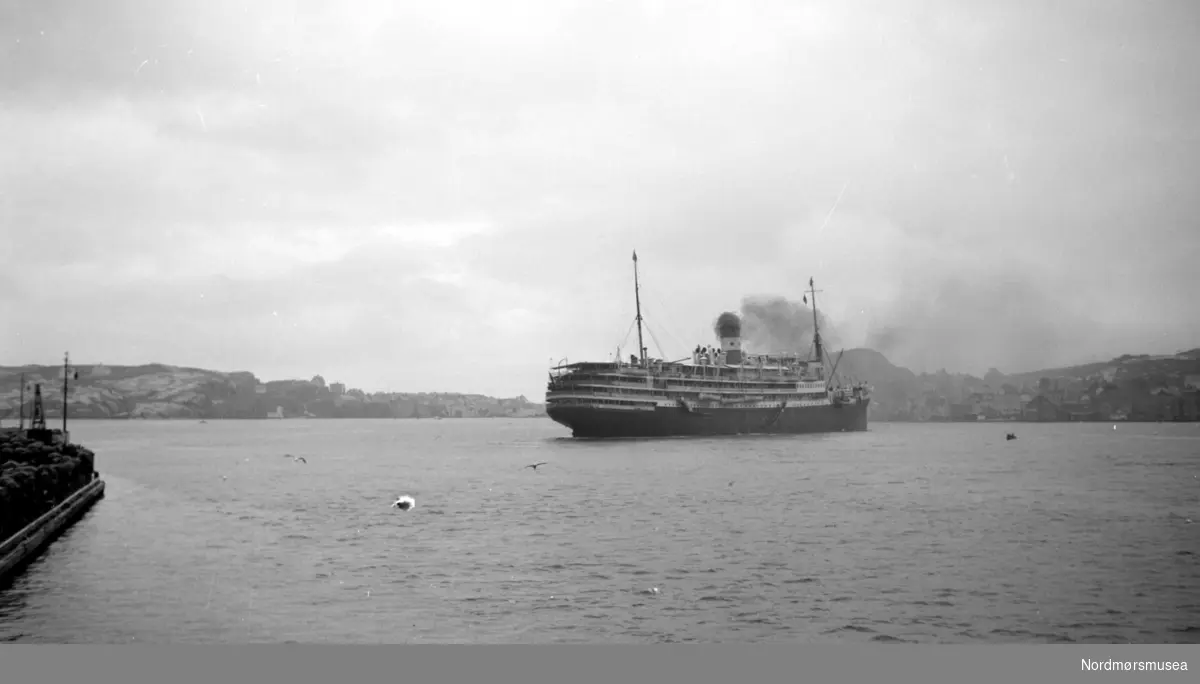 Turistbåten på bildet er franske M/S Foucauld på 11028 tonn, bygget for rederiet Compagnie des Chargeurs Reunis i 1923 som kombinert laste- og passasjerskip D/S L'Hoedic. Skipet ble ombygget til dieseldrevet turistskip i 1929 og gitt navnet M/S Foucauld.
Skipet gikk tapt 15.juni 1940 i La Pallice etter et tysk flyangrep. (Info fra Espen Arnestad 2017). Ute i havnebasenget i Kristiansund ser vi en passasjerbåt gå ut, med Gomalandet til venstre i bakgrunnen og Nordlandet i bakgrunnen til høyre.  Serie. Fra Nordmøre Museum sin fotosamling.