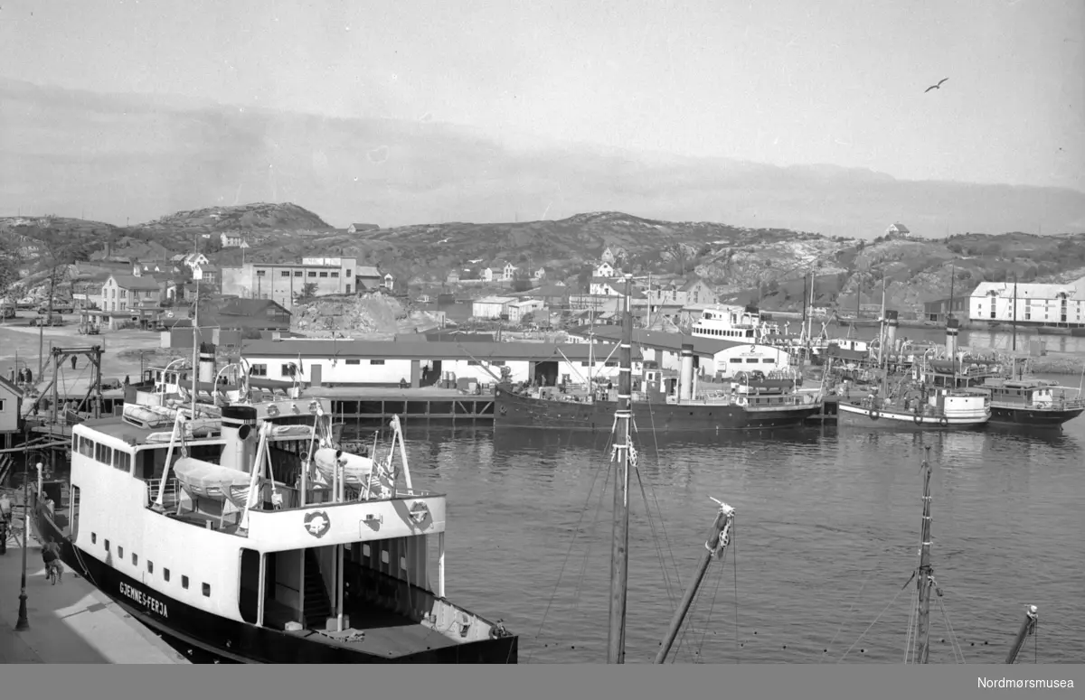 Foto fra gamle Johnsenhuken og Nordmørskaia ved havneområdet på Kirkelandet i Kristiansund, med "Gjemnes-ferja" i forgrunnen langs Vågekaia. Fra Nils Williams fotoarkiv. - "Gjemnes-ferja" hette egentlig B/F "Gjemnes". Det ble levert ny til Møre og Romsdal fylkesbåtar (MRF) i 1949 og satt inn i sambandet Kristiansund-Bremsnes-Kvernes-Bergsøy-Gjemnes-Torvikbukt. Sommeren 1952 var veien over Frei til Kvitnes ferdigbygd, og fergesambandet ble forkortet til Kvitnes-Bergsøy-Gjemnes-Torvikbukt. Men alt i 1954 ble B/F "Gjemnes" satt inn i sambandet Kristiansund-Bremsnes. 
Denne ferga gikk for MRF ca. 60 år. Den ble ombygd flere ganger og hette på slutten "Edøy". Kilde: Om samferdsel i Møre og Romsdal: Møre og Romsdal fylkesbåtar 1920-1995. (1995). [Molde]: Møre og Romsdal fylkesbåtar. (Info: Sveinung Berild)  - Fra Nordmøre museums fotosamlinger.