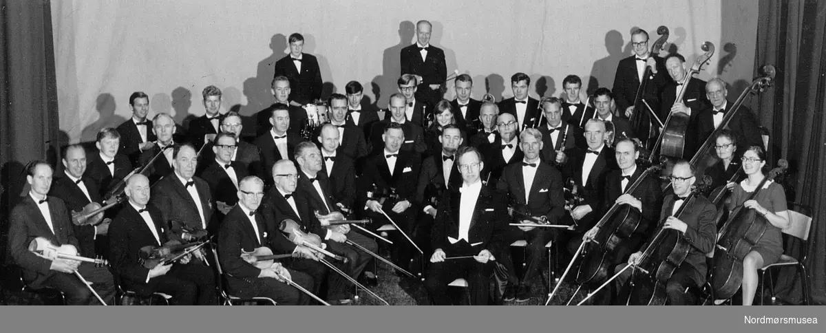 Kristiansund Symfoniorkester 50 år 1969. Dirigent Peder A Rensvik.

Rensviks første konsert som dirigent for KSO var 13.,15. og 16.mars 1969 i Festiviteten, på gymnaset og i Sunndal og Tingvoll. Harpesolist var Elisabeth Sønstevold, som Rikskonsertene også sendte til Ålesund på samme turen.  Fra denne produksjonen har vi bevart lydopptak av Schuberts ufullendte. Kristiansund formannskap fikk oversendt 34 billetter. Rensvik hadde stilt som forutsetning for å ta stillingen at kommunen opprettet musikkskole. Den fikk fra starten 1. september 150 elever, og lærerkreftene til de 80 i ”orkesterklassen” (herav 56 strykere) ble hentet fra orkesteret. Bokbinder Trygve Rossbach, selv utklekket på KSOs strykerskole av 1946, og baker Magnar Ersvik underviste begge på fiolin i 13 år. Frøydis Rensvik underviste på cello, Sigurd J. Nielsen underviste på obo, Ivar Olsen på fagott og Øivind Solbakk på horn. Igjen ble det appellert om at private måtte låne ut ledige fioliner.– 8.5.(Høgtun) og 9.5. (Festiviteten) var Kari Karlsen igjen solist, i Beethovens klaverkonsert nr.3 i C-moll. Otto Romfo og Arnstein Nilssen, nå politimester i Ålesund, var trompetsolister. Opptak ble gjort. – 25.10. stod KSO for musikken ved Odd Fellow-losje Runes 50-års-fest i Gripsalen og 13. og 15.11. var det 50-års-jubileum for KSO, på Tingvoll og i Festiviteten, med Mozarts 40.symfoni og prolog av Hedvig Wist. Det ble gjort lydopptak, og foto med navn står i Ohrviks hefte s. 33. Hindarkvartetten medvirket, i et samarbeid med Rikskonsertene og Molde Orkesterforening. De var i byen ei uke i oktober, for å instruere. Rikskonsertene og Norsk Kulturfond ga kr 17.500,- til dette. Kommunen 1500,- og staten 500,-. Kulturfondet ga også 4000 til et waldhorn. KSO tok opp et lån i Privatbanken på kr. 4100,- for å kjøpe sin første skikkelige kontrabass, en ¾ Pöllmann. Instrumentet ble kjøpt hos Theo Wang (Contronics forgjenger) og er i dag verdt 15 ganger mer. Kjøpet ble seinere refundert av Norsk Kulturfond. Rudolf Z. Unhjem ga KSO 35 notestativ i femtiårspresang. Fra kommunen ble det gitt ei skarptromme, og fra Musikkens Venner trommestativ og notestativ. Hermann Døhlen fra M.V. foreslo opprettet ”Edvard Bræins Minnefond” til støtte for orkestrets drift og utdanning av begavede musikere.  14.desember ble Mozarts Kroningsmesse framført i Bremsnes og Kirkelandet kirker. Averøy kommune ga et tilskudd på kr 300,-. – En bilutlodning ble forsøkt, men det ble kakelotteri på Rådhusplassen i stedet. Det innbrakte kr 1951,- over to lørdager.

Bilde fra Trygve Rossbachs dokumentasjon av Kristiansund Symfoniorkesters virksomhet på andre halvdel av 1900-tallet; ca 1950-2000. Prøver, turer, sosialt samvær og konserter. (Fra Normøre museum sin fotosamling.)