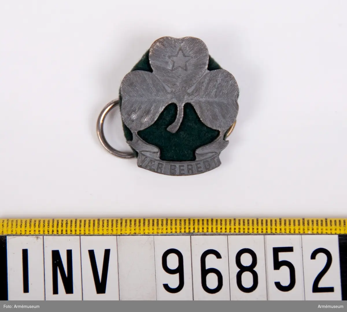 Ett märke i metall med formen av ett treklöver mot bakgrund av en liten filtbit i grönt tyg och på baksidan två metallringar fastsatta.