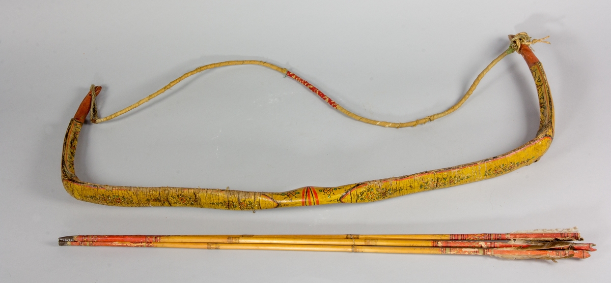 Pilbåge av trä. Böjd och avsmalnande på mitten. Två hak för att fästa strängen som är tillverkad av växtmaterial omlindat med bomullsband. Pilbågen gulmålad med blomsterdekor. Tre pilar av bambu delvis dekorerade i rött och svart. Metallspets och upptill fjädrar.