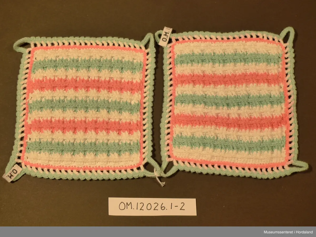 Firkanta grytelappar hekla i border med kvitt, rosa og lysgrønt garn. Rundt heile er det og ein heklakant i alle fargane, lysgrøn hempe i kvart hjørna.