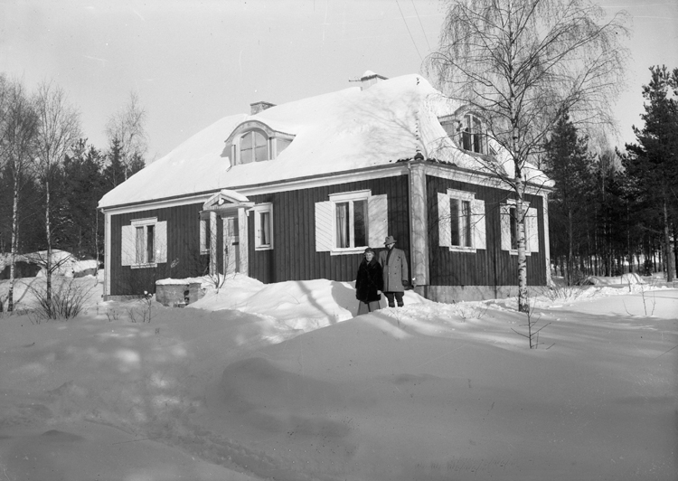 Foto av ett bostadshus. Vid ena knuten står en man och en kvinna. 
Vinterbild.