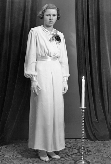 Foto av en ung kvinna i vit, lång konfirmationsklänning och vita handskar. Bredvid henne syns en golvljusstake med ett tänt ljus.
Helfigur. Ateljéfoto.