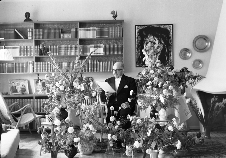 Foto av en äldre man i frack som sitter i ett vardagsrum, omgiven av blommor, och läser ett telegram. 
Kan ev. vara: Wictor Abraham Johansson (26 juli 1877-1960), Alvesta.
Källa: Sveriges Dödbok 1901-2009.