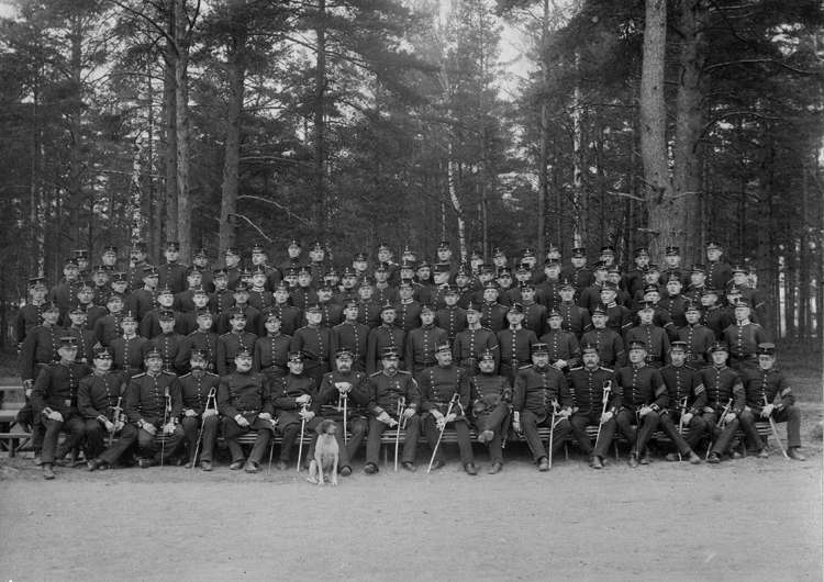 Gruppfoto av ett kompani med sina officerare (längst fram). 
De är fotograferade utomhus, mot en skogsdunge. Framför officersraden sitter en hund. 

Kronobergs hed (?).
