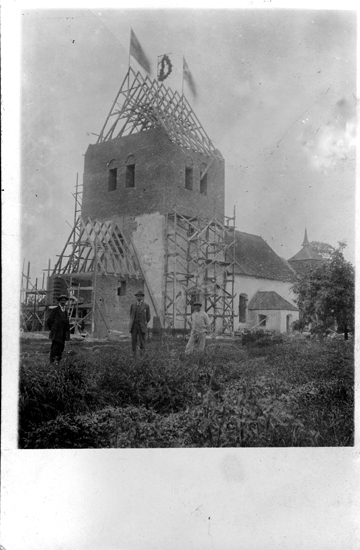 Foto på en kyrka som inte är färdigbyggd med två flaggor och en taklagskrans på tornet och två män som står bredvid. (Vykort).
