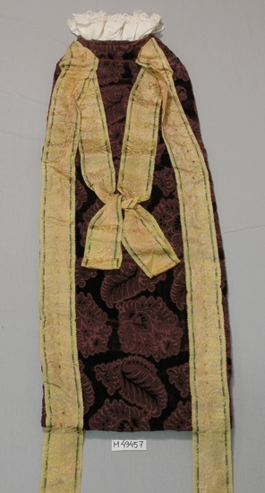 Dopdräkt av vinröd, mönstrad sammet.
Fodrad med rutigt tyg trol. lin i färgerna; gult, svart och ljusblått. I ovankant krage av vit bomull med knypplad 
spets.
Sprund bak (280 mm l) knytes med svarta ripsband. På framsidan sidenband (60 mm br) som dekoration
monterade efter långsidorna och som i sin förlängning blir "hängband" (450 mm l) i nedkant, i ovankant 
möts de i en rosett på mitten.
Sidenbanden är mönstervävda i färgerna; gult, rosa/beige och grönt.

Inskrivet i huvudbok 2007-08-16.
Funktion: Dopdräkt