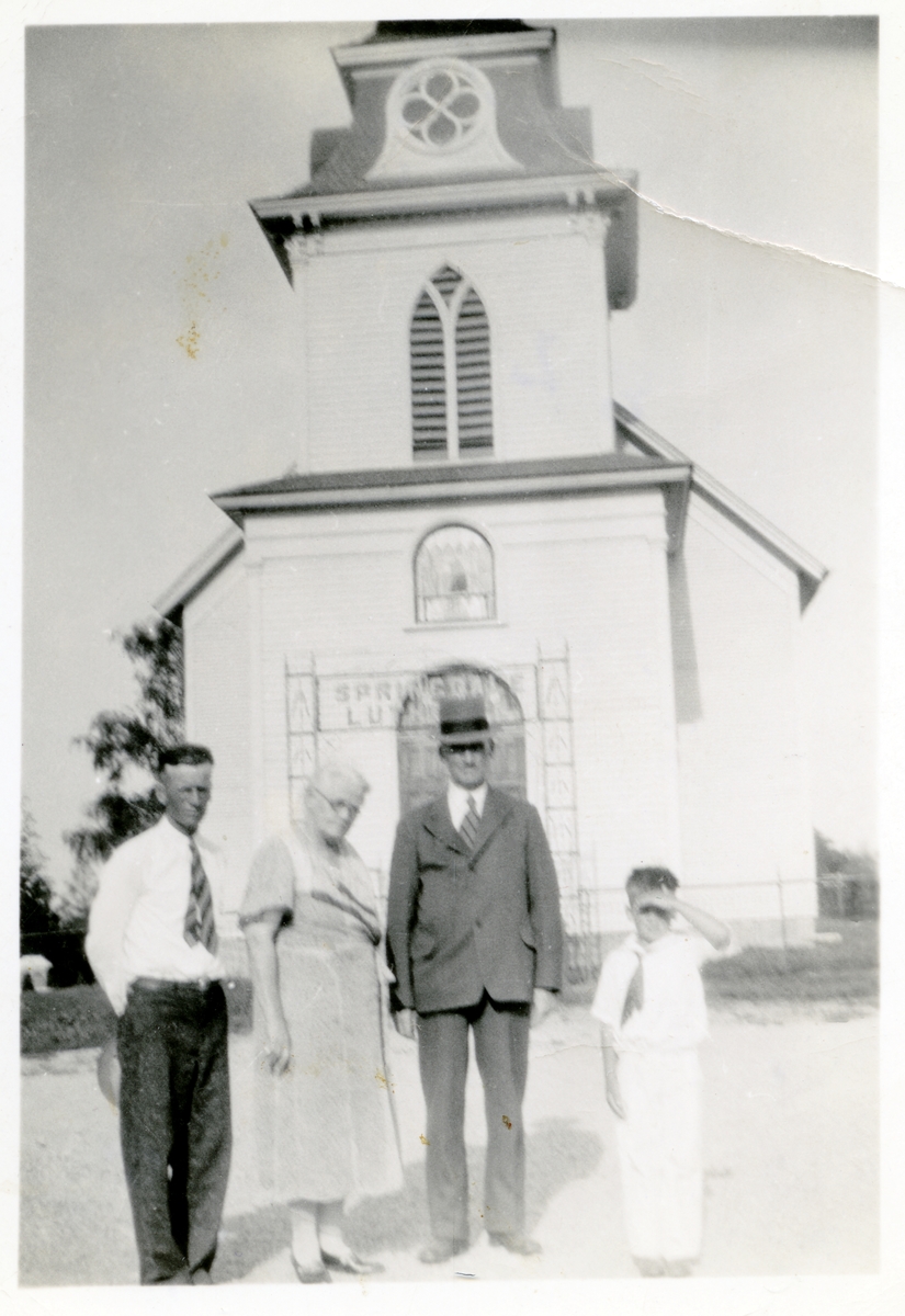 Springdale Lutheran Church i Wisconsin. En kvinne, to menn og en gutt står foran kirka. Mannen med dress og hatt er Olaus Islandsmoen.