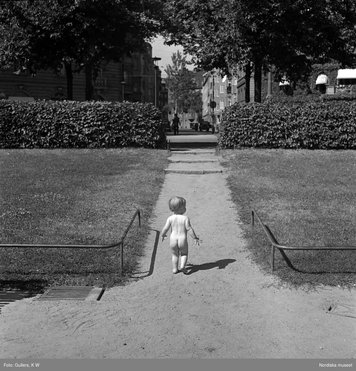 Ett litet naket barn promenerar på en liten grusväg utanför plaskdammen vid Karlaplan.