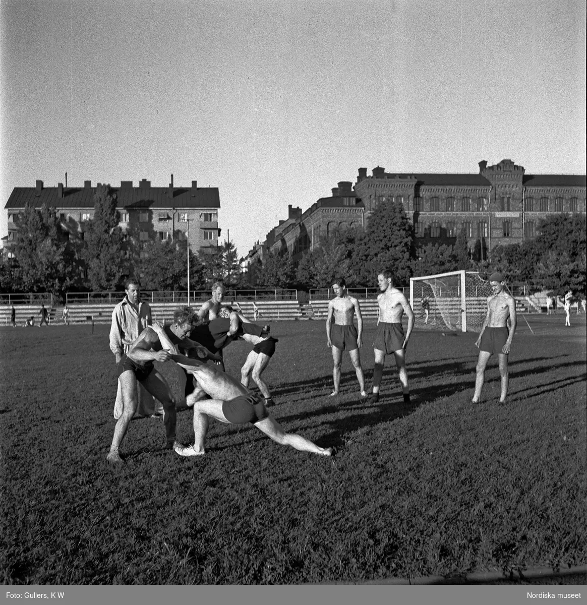 Zinkensdamms idrottsplats, Stockholm. Några pojkar brottas, andra pojkar som ser på. I bakgrunden ett nytt och ett gammalt hyreshus