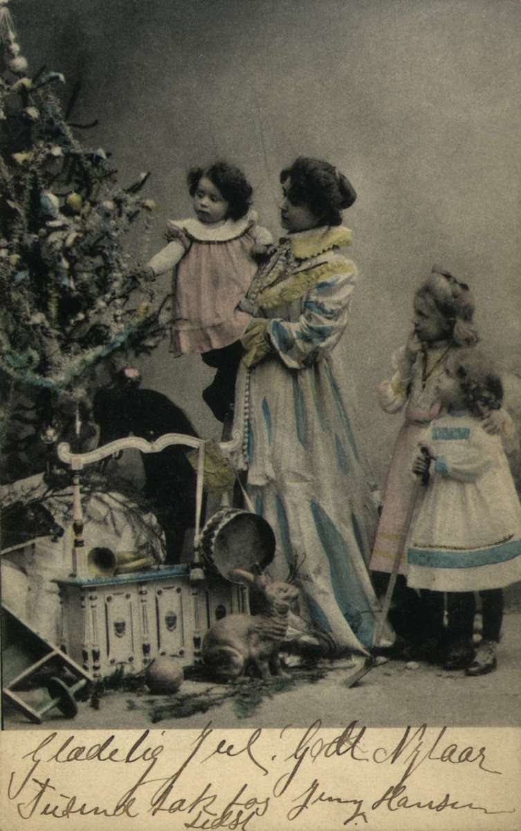 Julekort. Jule- og nyttårshilsen. Kolorert fotografi. Mor med tre barn ved pyntet juletre. Julegaver i forgrunnen. Stemplet 24.12.1904(?).