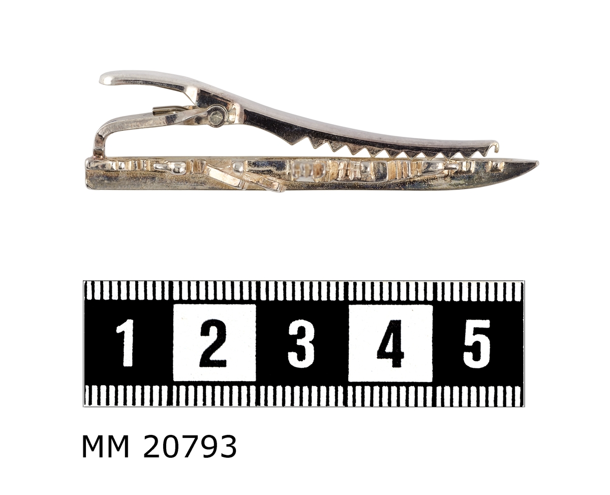 Silveraktig metall som föreställer en kustkorvett, Göteborgsklass, sedd från styrbordssidan. På baksidan av båten finns en tandad klämma. Under klämman är det två fördjupningar där det står "JOE" och "925".