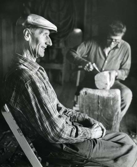 En leende man med arbetsbyxor, rutig skjorta och keps sitter med händerna i knät. Nan är modell åt skulptören Per Nilsson-Öst som här arbetar med ett stycke trä.