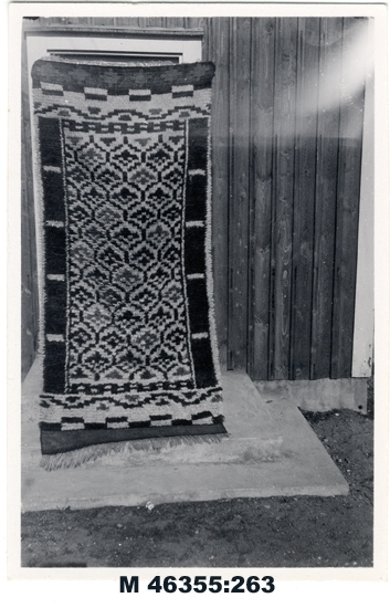 Svartvitt foto av flossamatta, utomhus.

Inskrivet i huvudbok 1983.
Montering/Ram: Ej ramad