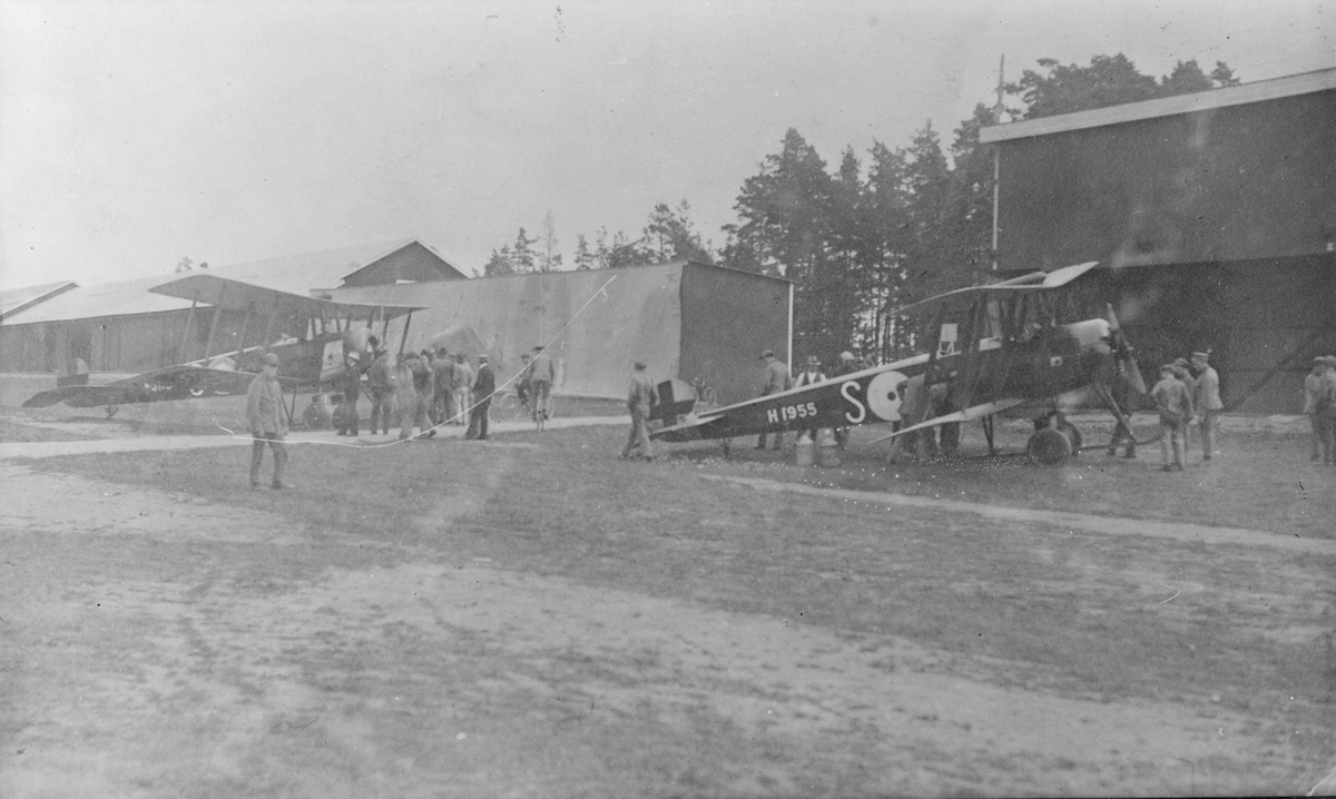 Människor i rörelse omkring två civila flygplan Avro 504K märkta 'S' utanför hangar på Centrala flygverkstäderna Malmslätt.