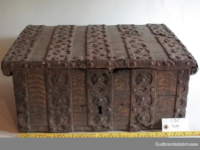 "Rik gjenombroten jarning over heile skrinet. Gamal type, alder uviss. Fra Vestre Gausdal". (Jf. "Katalog for Lom Bygdesamling,1925"). 
Skrinet har 3 rom, nøkkelhullslås og jernhandtak på sidene. Lokket er satt sammen av 2 bord, er festet til utsiden på bakstykket med to hengselbeslag og har 2 sidelister festet med trenagler. Bunnen er satt sammen av 2 bord, festet til hverandre med 2 forsenka trenagler; bunnen er festet til skrinrammen med trenagler.  Skrinrammen er tappet og forsterket med hjørnekantbeslag utvendig. 
Skrinet har jernbeslag med gjennombrutt mønster, 5 stk. B: 6,5 x L: 45 cm på lokk, 3 på hhv. bakstykke, front og sidestykker B: 6,5 x 20 cm. Lokket har ett låsbeslag og de to øvre hengselbeslag festet på innsiden, de to nedre hengselbeslag er festet på utsiden av bakstykket. Alle beslag er festet med store jernnagler samt festet under bunn på skrinet. De to hengselbeslag har også gjennombrutt mønster.  Nøkkelhullslåsen mangler låsanordning på innsiden.
DEKOR. Malingen har blitt beskyttet med et lag ferniss el.l., noe som ble vanlig fra begynnelsen av 1700-tallet. Ingen skurddekor. I stedet er beslag av jern brukt som dekorelement, noe som periodemessig kan peke på 1600-tallet, tidlig 1700-tallet, der treskurden ikke var fremherskende. Liknende dekorbruk finner du på LBY0328. Skrinet har gjennombrutt mønster på beslag og store jernnagler som beslagfeste. Treverket som synes mellom beslagene er malt med horisontale bølgestrukturer i gul og brun. Den dominerende dekorfargen er brun og gul, men det finnes små rester av blå eller blågrønn på lokk, sidelister på lokk, front og bunnkant på bakstykket samt av gul på frontbeslag. Innsiden av lokket er malt i terracottarød nyanse. Tradisjonelt fremkom denne fargen ved bruk av saft fra orr blandet med spytt, som ble smurt ut over treet. 
BRUK. Før kofferten kom i bruk hadde man reiseskrinet, som ble brukt til oppbevaring av bl.a. klær, verdisaker og mat på reiser. Skrinet hadde ofte et eller to mindre, smale rom langs den ene skrinsiden og ett større rom. De fleste reiseskrin var i tillegg forsterket med jernbeslag langs alle kanter og med en solid nøkkelhullås til sikring mot støt, slag og tyveri.