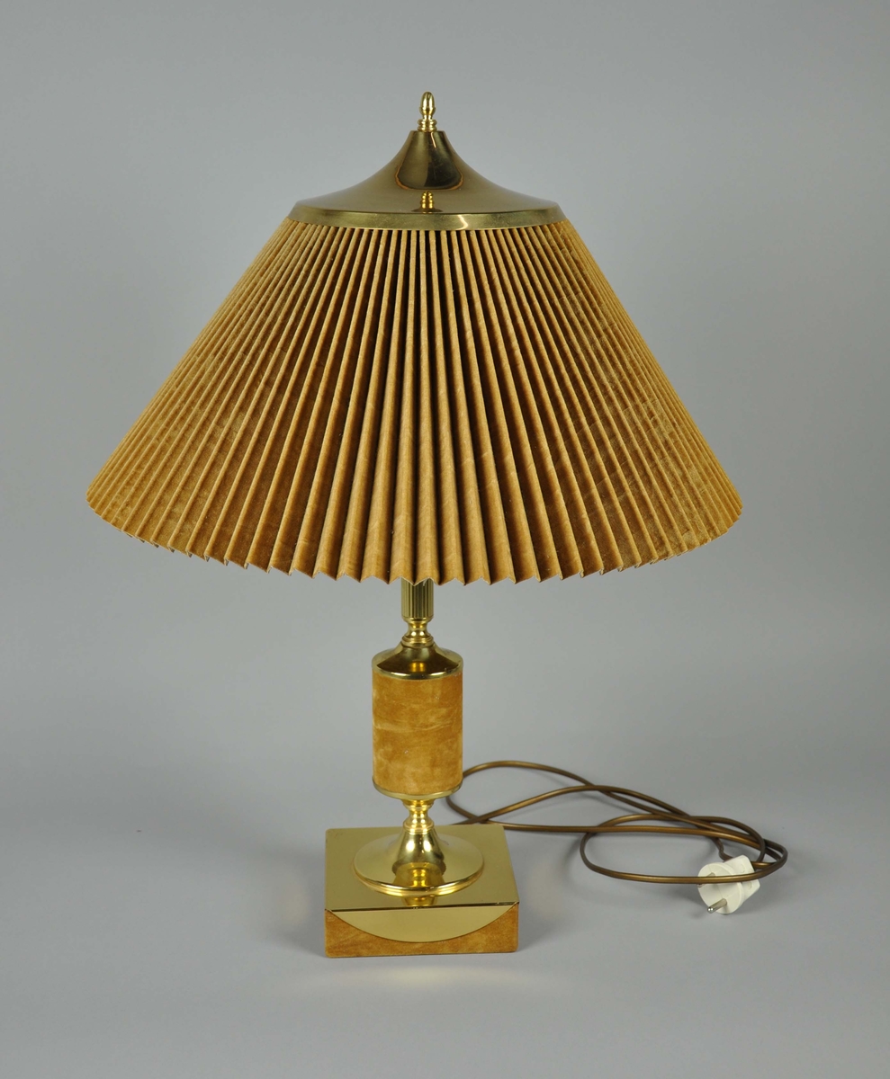 Lampe med kvadratisk fot og rillet øvre stett av messing. Midtre del av stetten er kledd med et  glatt og loddent stoff, trolig syntetisk. Skjermen er av det samme stoffet, er plissert og fôret med et plastmateriale.