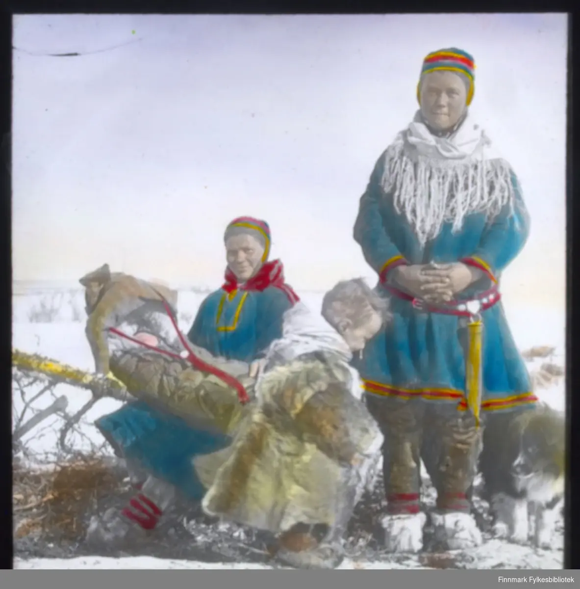 "N.549A9. Fjellsamer i Kautokeino" står det på glassnegativet. To samiske kvinner med sine barn er fotografert. Den ene sitter på en slede med et spebarn i komse. Barnet sover. Et litt større barn i pesk (reinsdyrpels) står foran i bildet. En samisk kvinne står oppreist. Begge er kledd i samisk kofte med sjal og lue. Kvinnen som står har en stor samekniv (storkniv) i beltet.