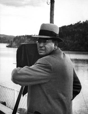 Svart-hvitt foto av fotograf med frakk, hatt og runde briller som titter til sida fra et kassefotoapparat.